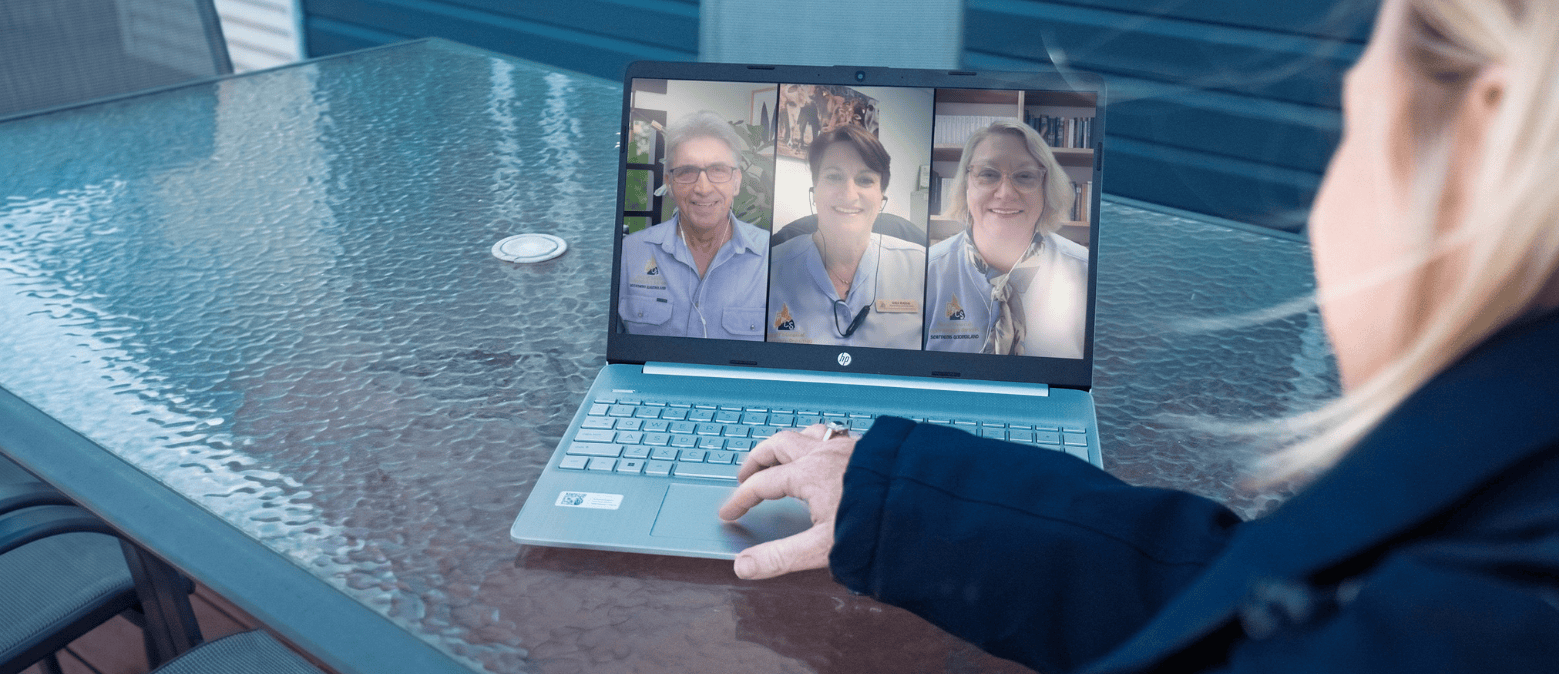 A lady is viewing a screengrab of webinar featuring a man and two women. The laptop is resting on a glass  outdoor table.